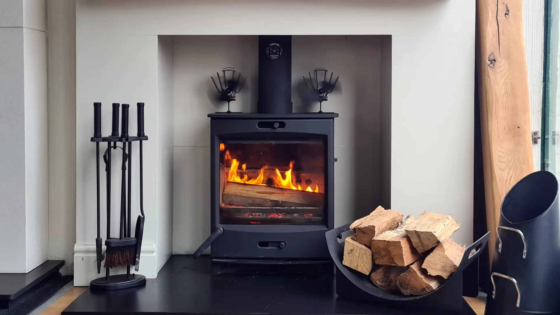 Black wood burning stove with logs ablaze, set in a white fireplace recess with fireside tools and a log holder